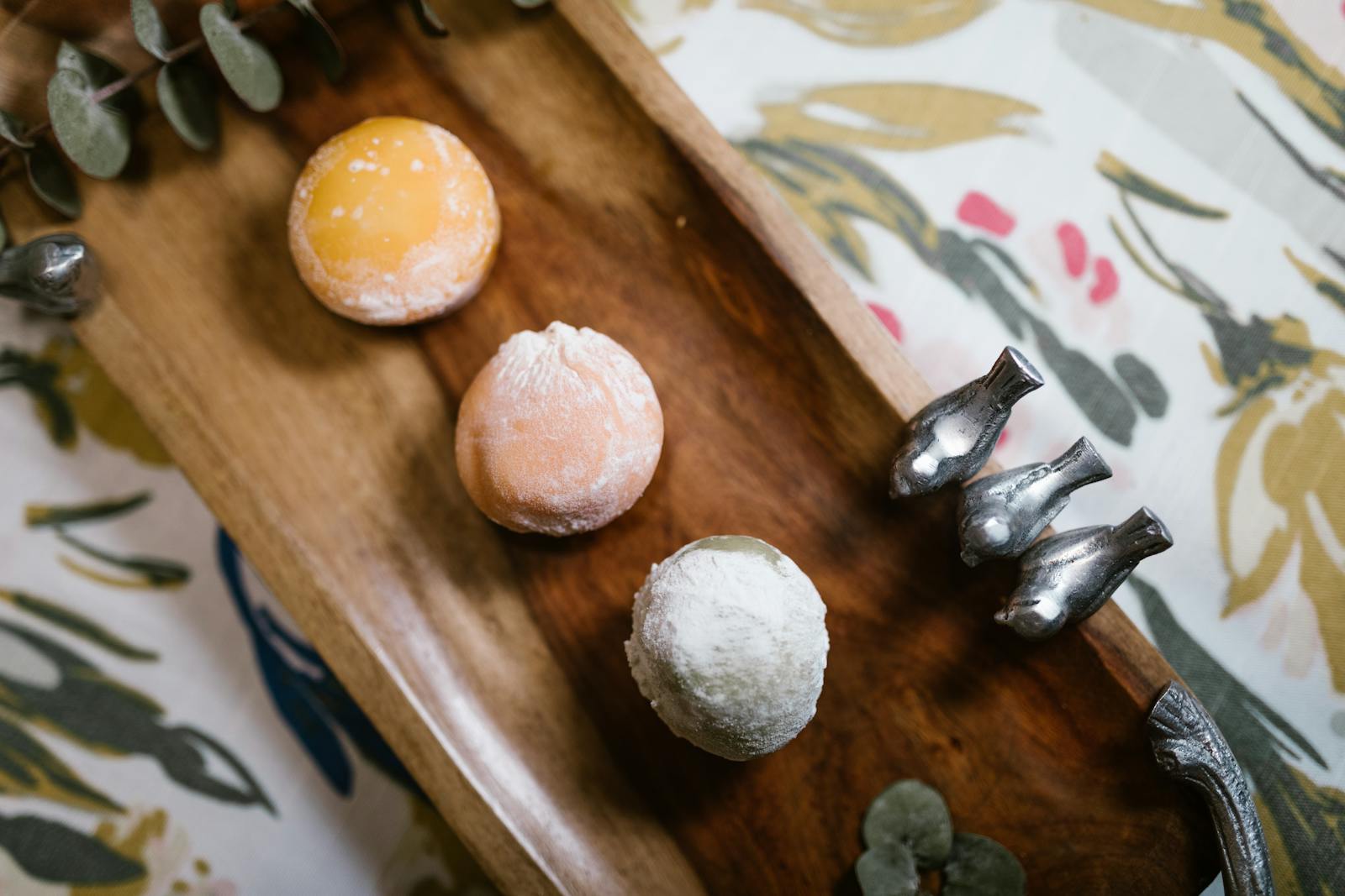 Tres mochi japoneses reales (amarillo, naranja claro, blanco) sobre tabla de madera oscura, con eucalipto y figuras de pájaros plateados al lado.